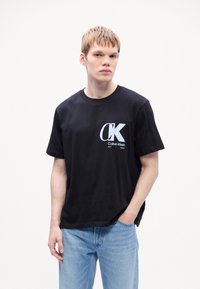 Musta puuvillapaita, jossa on valkoinen Calvin Klein -logo edessä. Pyöreä kaula-aukko, lyhyet hihat ja rento malli. Vaaleansiniset farkut.