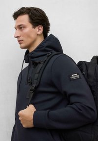 Sudadera con capucha de color azul oscuro y cremallera frontal, con un parche en la manga. El modelo lleva una mochila negra con correas ajustables.