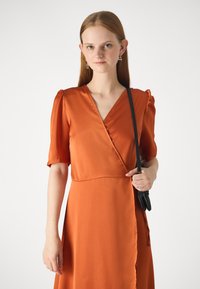 Narančasta haljina s kratkim napuhanim rukavima, glatke teksture i V-izrezom. Model drži crnu torbicu, ističući elegantan draperiju haljine.