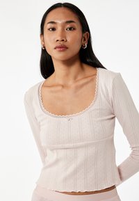 Ljusrosa långärmad topp med rundad halsringning, strukturerat ribbat tyg och ömtålig vågig kant längs kanterna. Diskret prickmönster.