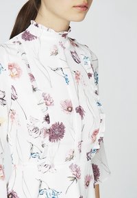 Blommig klänning med vit bakgrund, prydd med rosa och blå blommor. Rynkad halsringning och detaljer längs fram och ärmar.