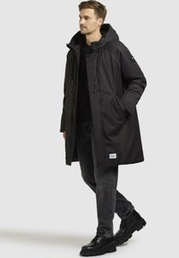 Sort isoleret parka med hætte, lynlås- og knaplukning, sidelommer og justerbare snore; parret med mørkeblå jeans og sorte støvler.