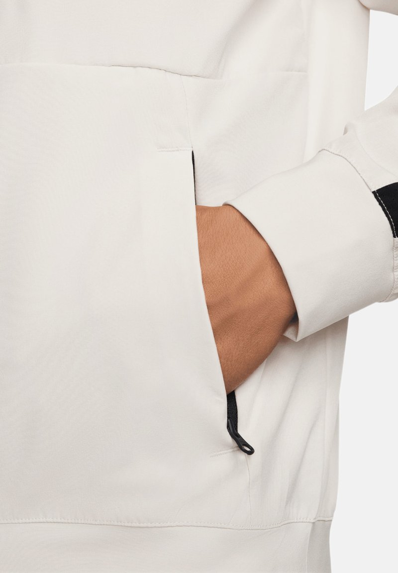 Chaqueta ligera blanca con un bolsillo con cremallera. La manga presenta una franja de acento negra. Textura suave y diseño limpio.