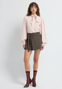 Blusa en rosa claro con un lazo, combinada con una mini falda envolvente marrón que presenta un detalle de hebilla. Medias transparentes burdeos y tacones completan el conjunto.