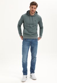 Man in een groene hoodie, blauwe jeans en witte sneakers, staande met zijn handen in de zakken tegen een effen achtergrond.