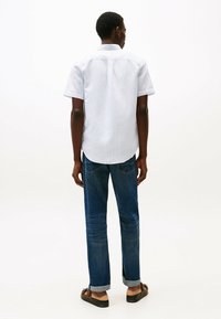 Camisa de manga corta y botones en azul claro con un sutil patrón de rayas, combinada con jeans azul oscuro y sandalias marrones. Posicionada desde detrás.