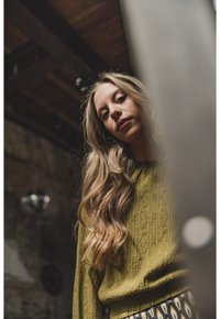 Olijfgroene kabelgebreide trui met lange mouwen, gecombineerd met een patronenrok. Lang, golvend haar bedekt gedeeltelijk het gezicht.