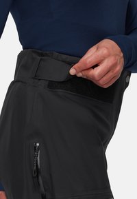 Zwarte waterdichte broek met een klittenbandsluiting en een ritszak aan de zijkant. De stof lijkt glad en duurzaam, ontworpen voor gebruik buitenshuis.