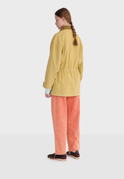 Donna con i capelli intrecciati vista di profilo verso sinistra, indossa una giacca giallo senape, pantaloni rosa dalla trama strutturata e scarpe marrone scuro su uno sfondo bianco.