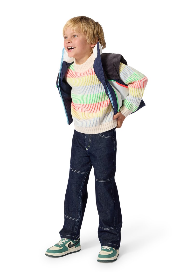 Jeune garçon portant un pull rayé coloré, un jean foncé, des baskets vertes et blanches, enfilant une veste bleu marine, souriant et regardant vers le haut.