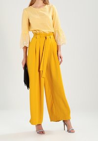 Gula vida byxor med en knytbälte, i kombination med en texturerad gul topp med spets ärmar i klockform. Svart kuvertväska och blå klackar.