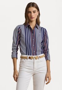 Camicia a righe blu navy e bianca con accenti rossi. Presenta un colletto e maniche arrotolate, abbinata a pantaloni bianchi e una cintura di corda.
