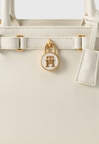 Λευκή δερμάτινη τσάντα με ανάγλυφη υφή, διακοσμημένη με μεταλλικά στοιχεία σε χρυσό τόνο, συμπεριλαμβανομένης της κλειδαριάς με λογότυπο και μιας μικρής ετικέτας. Καθαρά γραμμές και δομημένος σχεδιασμός.