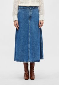 Džinsa maksi svārki vidēji zilā krāsā ar priekšējo šuvi, augstu jostasvietu un nedaudz izplatu apakšu, savienoti ar brūnām patentādas zābakiem.