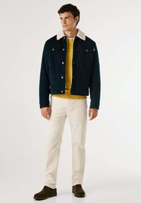 Giacca di velluto a coste blu con colletto in shearling bianco, abbinata a un maglione giallo e pantaloni beige. Stivali alla caviglia in suede marrone.