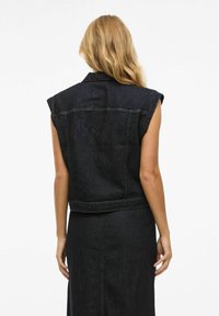 Mörk denim ärmlös jacka med axelvaddar, rå fåll och synliga sömnadsdetaljer. Slät textur, åtsittande design och avslappnad stil.