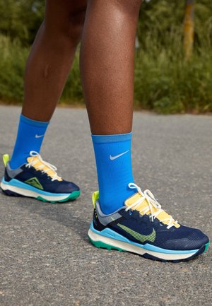 Ben iført lyseblå Nike-strømper og marineblå, gule og grønne Nike-løbesko stående på asfalt udendørs.