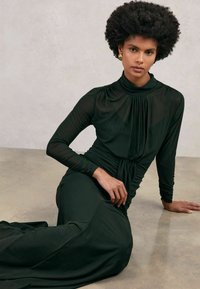 Pikk tume roheline kleit kõrge kaelusega, pikkade läbipaistvate varrukatega ning kogumisega detailide külgedel, asetatud neutraalsele pinnale.