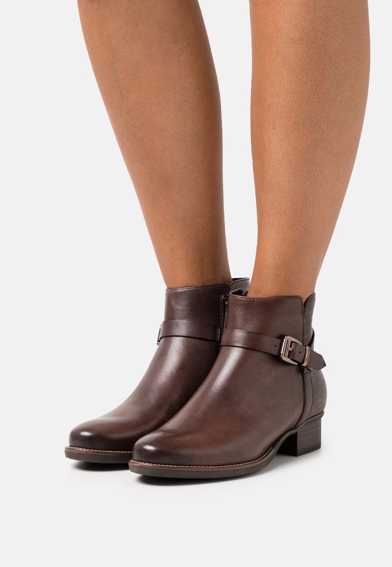 Bottes basses en cuir marron avec des brides à boucle sur le côté, portées sur des jambes nues sur un fond blanc uni.