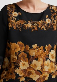 Blusa floral con base negra y patrones de pensamientos y flores en amarillo dorado, cuello redondo y mangas largas. Textura de tela suave.
