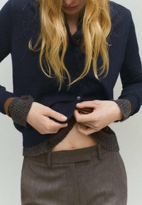 Námornícky kardigán s gombíkovou prednou časťou, hnedými rebrovanými manžetami a príjemným šedým lemom, vrstvený cez šedé nohavice, odhaľujúci strednú časť brucha.