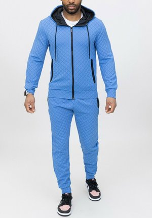 Ensemble de survêtement bleu matelassé avec une veste à capuche zippée et des cordons, comportant des accents noirs et des poches latérales, associé à des baskets noires.