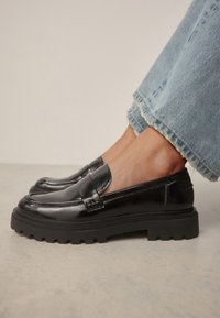 Black patent leather loafers with a chunky rubber sole and rounded toe, featuring a stitched vamp and smooth surface texture.