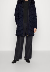 Námořnická péřová bunda s kapucí lemovanou kožešinou, zapínáním na zip a proužkovým prošíváním ve tvaru chevronu, nošená přes tmavé tričko a široké džínové kalhoty.