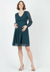 Tiefgrünes Umstandskleid mit V-Ausschnitt, langen durchsichtigen Ärmeln, plissiertem Rock und tailliertem Bund. Kombiniert mit Riemchen-Pumps.