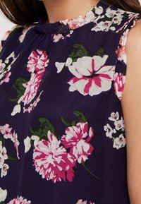 Květinová bezrukávová blůza s tmavě purpurovým základem, velkými růžovými a bílými květinami a volánkovým výstřihem. Měkká a lehká tkanina.