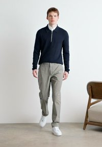 Marineblå genser med glidelås, lysegrå bukser og hvite sneakers. Modellen står på et beige gulv med en trestol i nærheten.