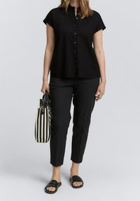Camicia nera a maniche corte con bottoni, abbinata a pantaloni neri slim-fit e sandali neri a scivolo. Il modello tiene una borsa a tracolla a righe.