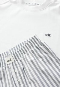 Bijela pamučna majica s izvezenim logotipom "self.", uparena sa sivim i bijelim prugastim pamučnim kratkim hlačama za spavanje s elastičnim pojasom i gumbima.