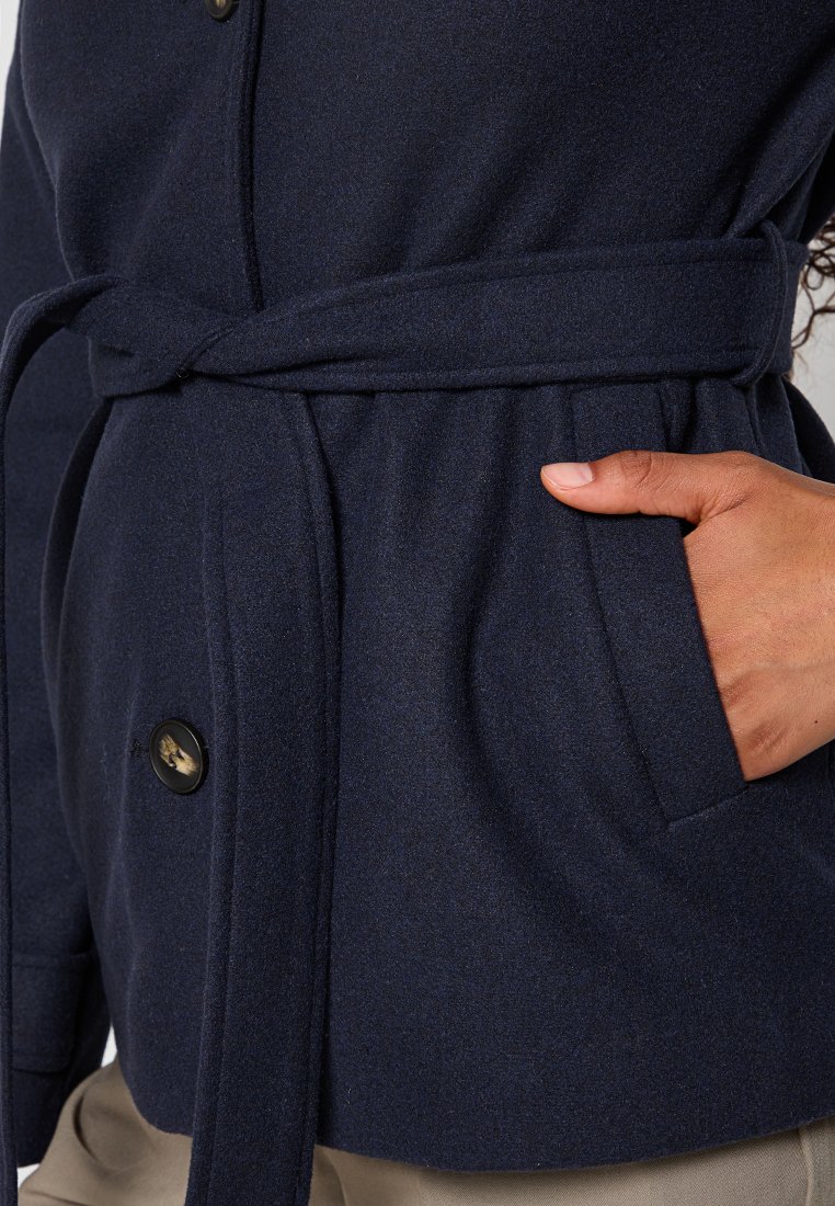 Manteau texturé bleu marine avec une taille ceinturée, doté de boutons foncés et d'une poche latérale. Le tissu semble doux et chaud.