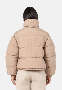 Beige Steppjacke mit hohem Kragen, gestepptem Material und elastischen Bündchen. Hat ein cropped Design und eine lockere Passform.