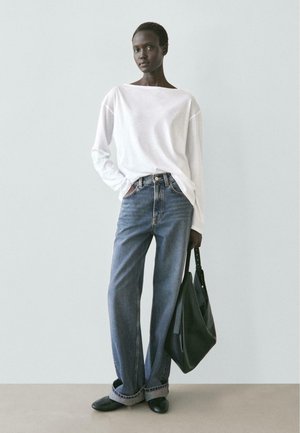 Donna che indossa una camicia bianca larga a maniche lunghe, jeans blu risvoltati, scarpe nere e tiene una grande borsa nera davanti a uno sfondo neutro.