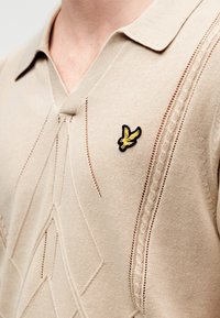 Μπεζ πλεκτό πόλο με V-λαιμόκοψη, υφασμένα σχέδια και διακοσμητικό μαύρο και κίτρινο κεντητό λογότυπο πουλιού στο στήθος.