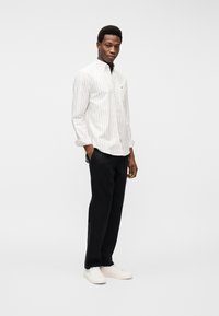 Άνδρας στέκεται με τα χέρια στις τσέπες, φοράει λευκό ριγέ πουκάμισο, μαύρο παντελόνι και λευκά αθλητικά παπούτσια, μπροστά σε απλό φόντο.