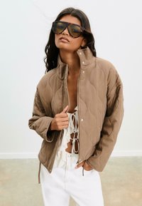Femme portant des lunettes de soleil surdimensionnées en écaille de tortue, une veste matelassée beige partiellement zippée, et un pantalon blanc avec un top blanc à volants noué à l'avant.