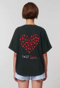 Osoba s krátkými vlasy nosí tmavé tričko, na kterém je velké červené srdce ze menších srdcí a pod ním je text "SWEET heart".