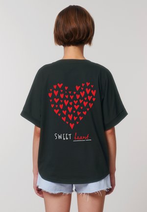 Une personne avec les cheveux courts porte un t-shirt noir avec un grand cœur rouge composé de cœurs plus petits et le texte "SWEET heart" en dessous.
