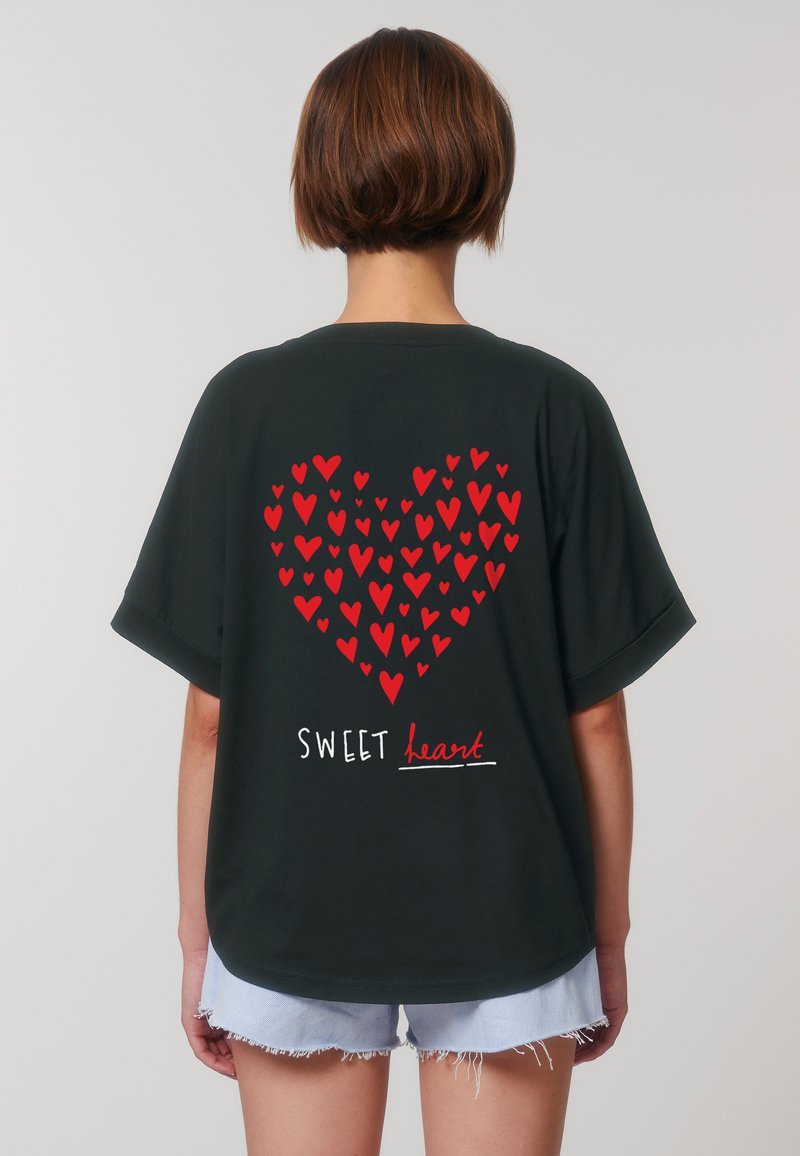 Osoba s krátkými vlasy nosí tmavé tričko, na kterém je velké červené srdce ze menších srdcí a pod ním je text "SWEET heart".