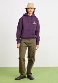 Purple hooded sweatshirt with a front pocket and triangle logo, paired with light brown trousers and black slip-on shoes, worn with a beige bucket hat.