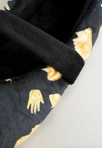 Zapatos de tela negra con patrones de manos y conchas amarillas, que cuentan con una textura suave y un diseño aerodinámico. Incluyen una correa negra en la parte superior.