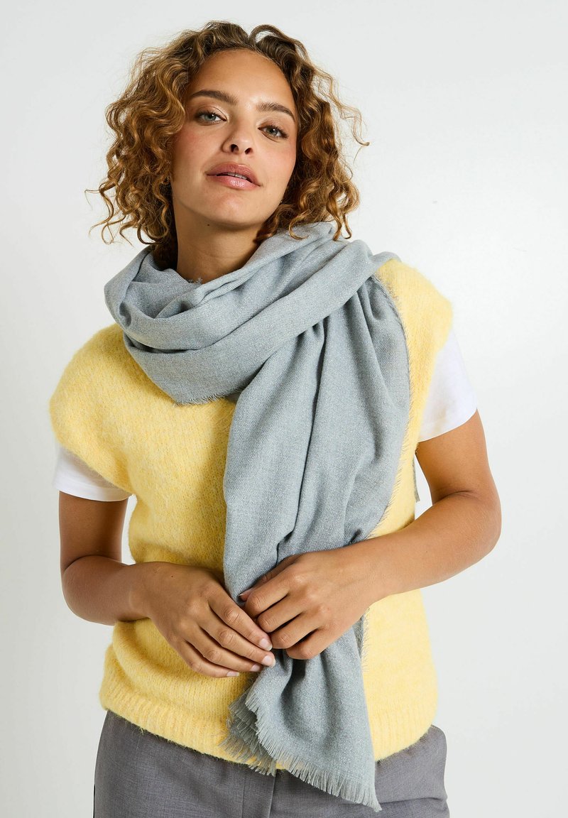 Bufanda azul clara hecha de una tela suave y texturizada, colocada alrededor del cuello, con bordes con flecos; combinada con una blusa amarilla de felpa y pantalones grises.