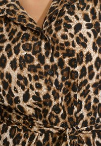 Blúzka s leopardím vzorom, ktorá má gombíkový uzáver, čierne akcenty a viazaný pás. Látka má hladkú, jemne lesklú textúru. Farby sú hnedá, čierna a biela.