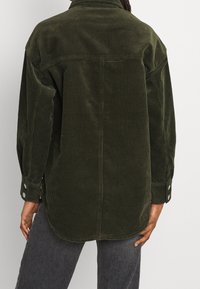 Tmavě zelená košile z manšestru s volným střihem, zaobleným lemem a manžetami na knoflíky, s vertikální žebrovanou texturou a límečkem.