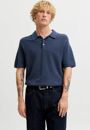 Jeune homme aux cheveux blonds bouclés portant un polo en maille bleu marine à manches courtes et un jean foncé avec des tatouages sur les deux avant-bras.
