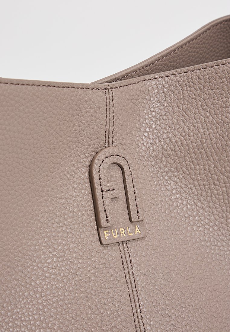 Beżowa skórzana torba o drobnej teksturze, z przeszywaną szwem i złotą metką z logo. Minimalistyczny design, otwarta górna część.