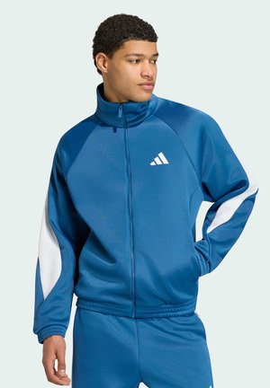 Giovane uomo che indossa una tuta Adidas blu con strisce bianche, in piedi con una mano in tasca, che guarda di lato.